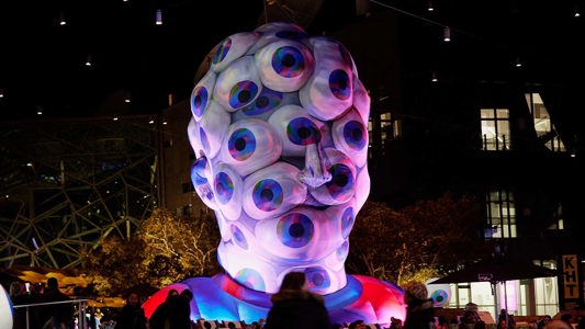 A large inflatable structure in the shape of a human head that is coloured purple and silver is covered in eyes looking outwards, with purple light all around.