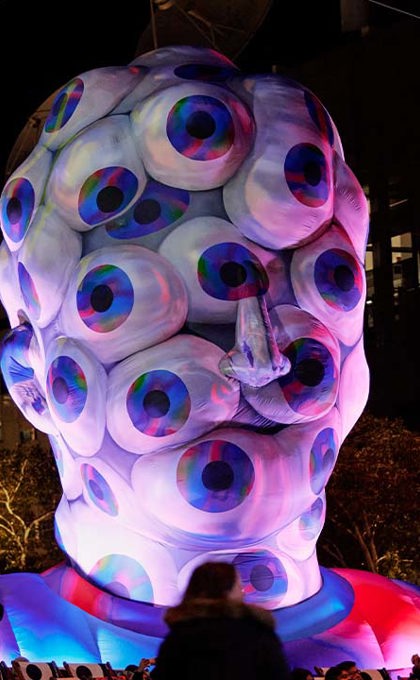 A large inflatable structure in the shape of a human head that is coloured purple and silver is covered in eyes looking outwards, with purple light all around.